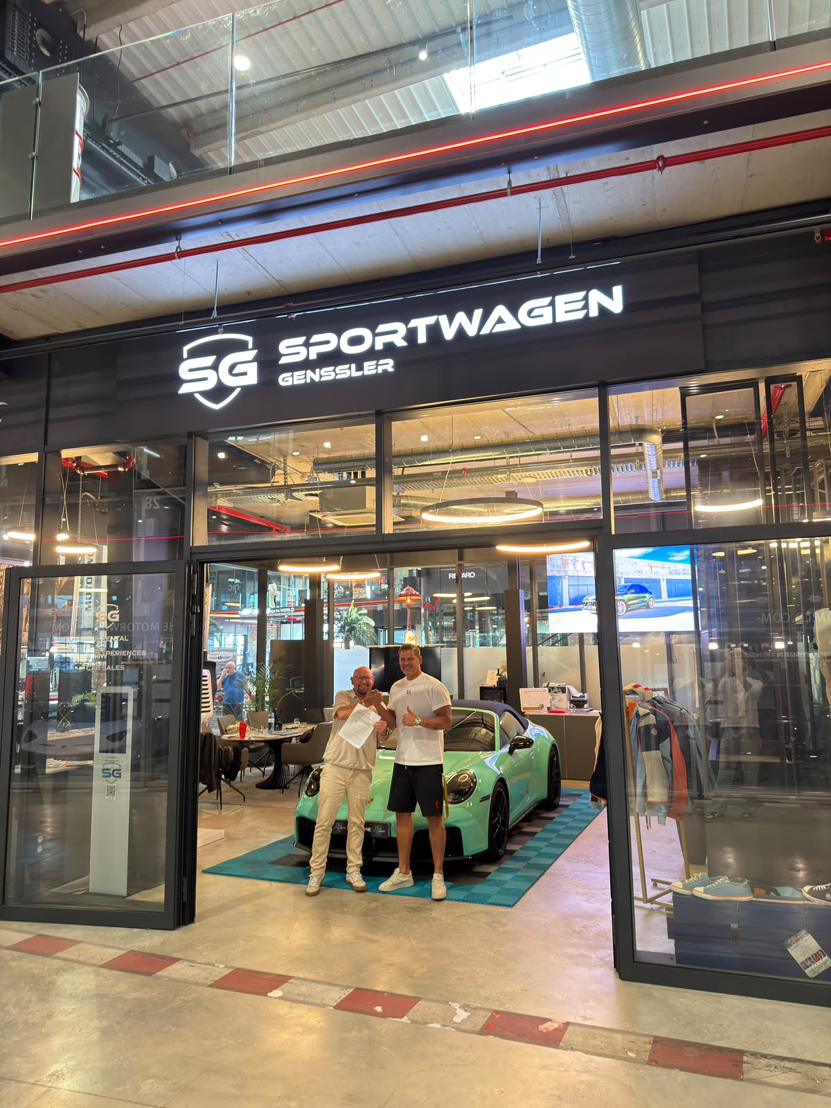 Gassi hält vor dem Showroom von SG Sportwagen Genssler in der Motorworld Palma den unterschriebenen Vertrag hoch, dahinter ein mintgrüner Porsche 911